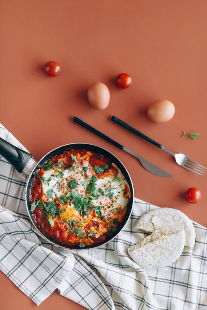 Pan of eggs baked in tomato sauce with fresh herbs, flatbread on a checked cloth