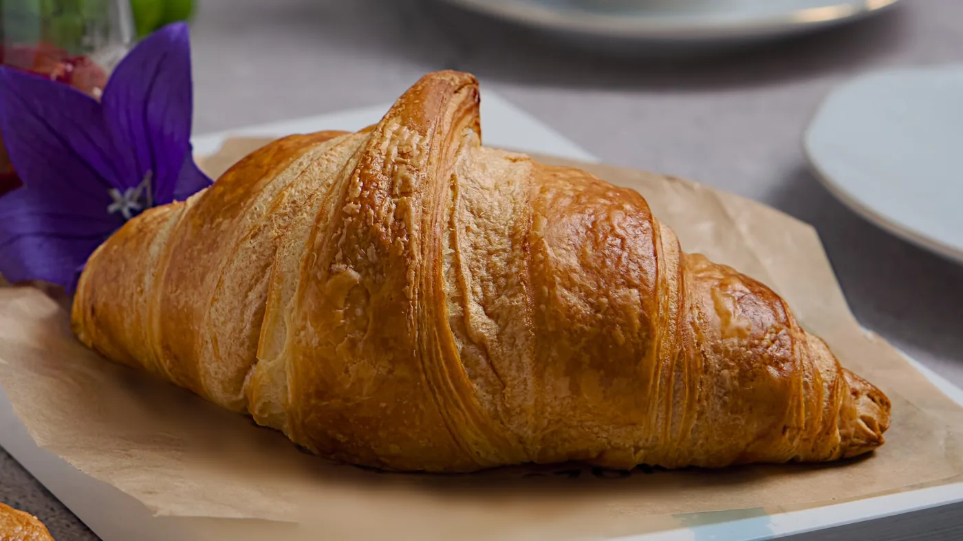 Croissant on brown paper, beside a purple flower, with a cup and saucer behind